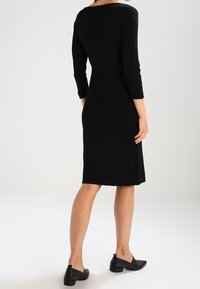 Robe noire moulante à manches trois-quarts et col rond. Texture lisse, longueur au genou, portée avec des chaussures plates noires.