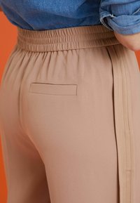 Pantalon beige avec une taille élastique, des rayures latérales et une petite poche arrière. Tissu lisse avec une texture douce.