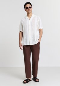 Chemise blanche à manches courtes avec un motif de tricot ouvert, boutons jaunes ; pantalon droit marron ; sandales noires avec un design perforé.