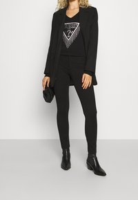 Svart blazer över en svart grafisk topp med silverstrass. Matchande leggings och ankelskor fulländar outfiten. Enkel, monokrom design.