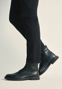 Czarne skórzane botki za kostkę z sznurówkami i suwakami po bokach, noszone z czarnymi dopasowanymi spodniami na jednolitym jasnym tle.