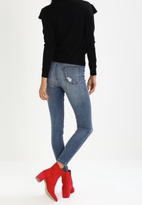 Top noir à manches longues, jeans skinny déchirés bleu clair et bottines rouges à talons carrés, vus de derrière sur un fond neutre.