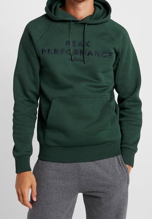 Mann trägt einen dunkelgrünen Hoodie mit der Aufschrift "PEAK PERFORMANCE ORIGINAL" und graue Jogginghose, steht mit einer Hand in der Tasche.