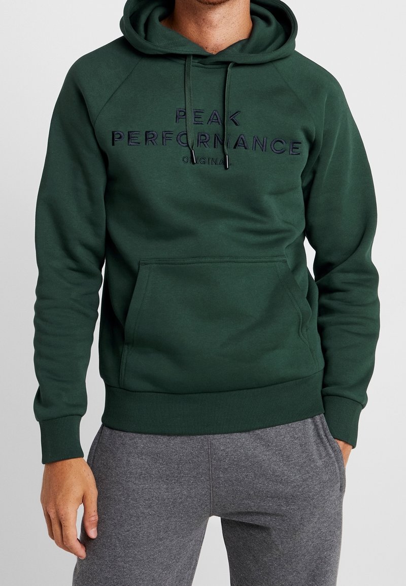 Homme portant un sweat à capuche vert foncé avec le texte "PEAK PERFORMANCE ORIGINAL" et un pantalon de survêtement gris, debout avec une main dans la poche.