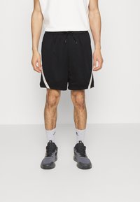 Schwarze Sportshorts mit Netzstruktur, versehen mit weißen Seitenakzenten, einem elastischen Bund mit Kordelzug. Getragen mit dunklen Turnschuhen und grauen Socken.