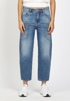 Woman wearing light blue straight-leg jeans with large front pockets and white sneakers standing against a plain background.