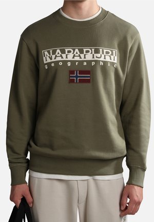 Zielona bluza z napisem "Napapijri geographic" i naszywką norweskiej flagi na piersi, noszona na białej koszuli z beżowymi spodniami.
