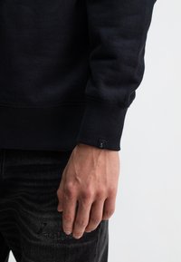 Zwarte sweatshirt met geribbelde manchetten en een klein merkje aan de mouw. De stof lijkt zacht; het ontwerp is eenvoudig en ongecompliceerd.