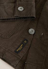 Hnedá tkanina s textúrovaným povrchom, s pevním strieborným gombíkom a štítkom s nápisom "Durable Goods" s žltými akcentmi.