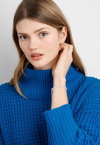 Blue knitted sweater with a high collar design and textured pattern. Model touches her neck, wearing a silver bracelet.
