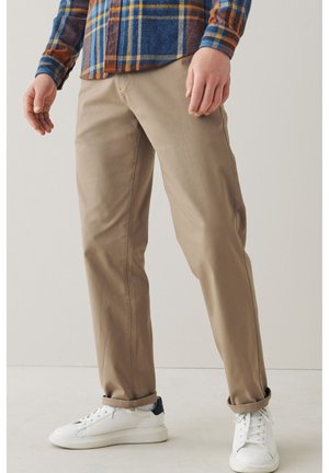 Chinos beige con un corte recto, hechos de tela suave. Llevados con zapatillas blancas con un acento azul marino y presentados sobre un fondo claro.