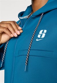 Teal zip-up hoodie made of smooth fabric, featuring a front pocket, black and white drawstrings, and a white logo on the chest.