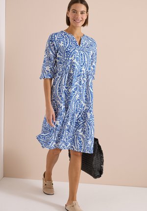 Kobieta idąca w niebiesko-białej kwiecistej sukience do kolan, trzymająca czarną plecioną torbę i nosząca beżowe buty wsuwane.