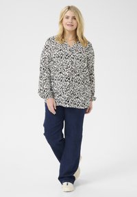 Schwarz-weiß gemusterte Bluse mit Kragen und langen Ärmeln, kombiniert mit dunkelblauen geraden Jeans und hellen Sneakers.