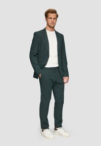 Grüner Anzug aus strukturierter Qualität, einreihige Blazerjacke, passende Hose, kombiniert mit einem weißen T-Shirt und weißen Sneakers.