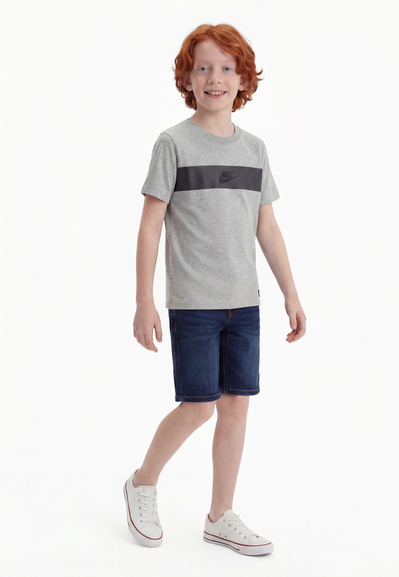 Vörös hajú fiú, szürke Nike pólóban, sötét farmer rövidnadrágban és fehér tornacipőben, fehér háttér előtt állva mosolyog.