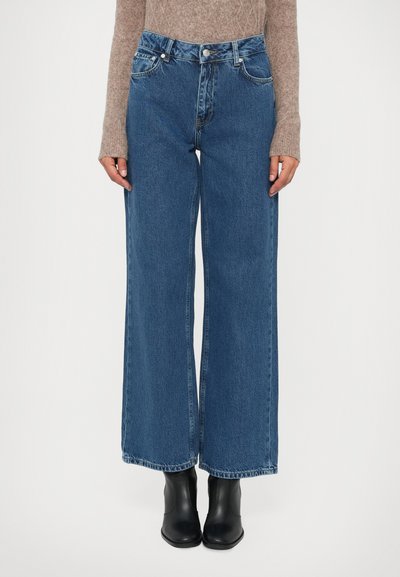 Vrouw in hoge taille, wijde blauwe jeans, beige lange mouwen trui en zwarte enkellaarsjes, staande tegen een effen achtergrond.