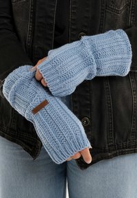 Lichtblauwe gebreide vingerloze handschoenen met ribbelstructuur en een leren accentlabel, gedragen met een zwarte jas en een spijkerbroek.