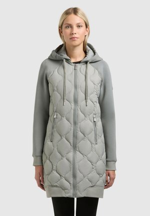 Femeie purtând o jachetă lungă matlasată de culoare gri deschis, cu glugă, fermoar frontal și buzunare cu fermoar, stând în fața unui fundal simplu.