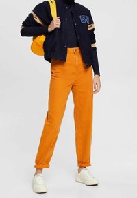 Marineblaue Bomberjacke mit beigen Akzenten, schwarzer Rollkragenpullover, orangefarbene, hochtaillierte Hose und weiße Turnschuhe; lässiger Streetwear-Look.