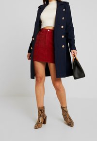 Frau trägt einen marineblauen, doppelt gelegten Mantel, ein weißes Crop-Top, einen roten Cordrock und Schlangenleder-Print Stiefeletten, während sie eine schwarze Handtasche mit Holzgriffen hält.