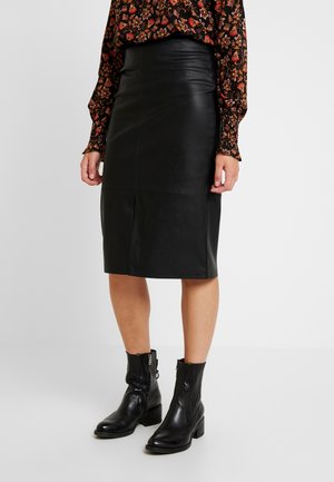 Femme portant une jupe crayon en cuir noire longueur genou, des bottines noires à talons, et un chemisier à manches longues à motif floral, debout.