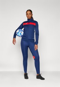 Giacca sportiva blu navy con accenti rossi, chiusura con zip e strisce bianche, abbinata a leggins coordinati con una toppa rossa e scarpe nere.