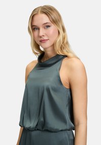 Cowl-neck top in metallic teal fabric with a soft, textured finish and sleeveless design, featuring a loose fit and gathered waistband.