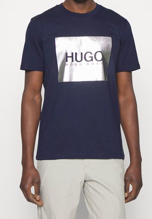 Homme portant un T-shirt bleu marine à manches courtes avec un carré argenté brillant et le texte "HUGO HUGO BOSS" sur la poitrine, associé à un pantalon beige.