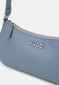 Niebieska skórzana torebka o fakturze groszkowej, z logo "HUGO" w kolorze srebrnym i zamknięciem na zamek błyskawiczny na górze. Posiada odpinany pasek, co umożliwia różne sposoby noszenia.