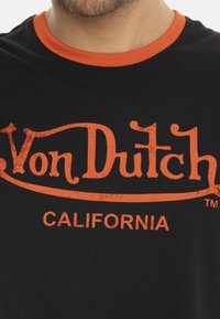 T-shirt noir avec un col rond orange et un texte orange usé affichant en grande taille "Von Dutch CALIFORNIA" sur le devant.