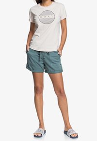 Světlobéžové tričko s logem Roxy, zelenomodré cargo kraťasy s ohrnutými lemy a modré nazouvací sandály s pruhovaným vzorem.