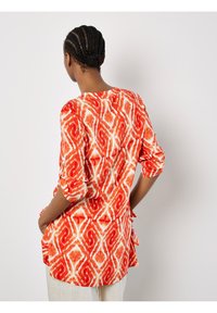 Rot-weiß gemusterte Bluse mit langen Ärmeln, Rundhalsausschnitt und seitlichen Schlitzen für Bewegungsfreiheit. Leichtes, strukturiertes Material.