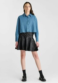 Geknipte blauwe denimshirt met een knopenfront, gecombineerd met een zwarte faux leren flared rok. Het model draagt zwarte sokken en schoenen.