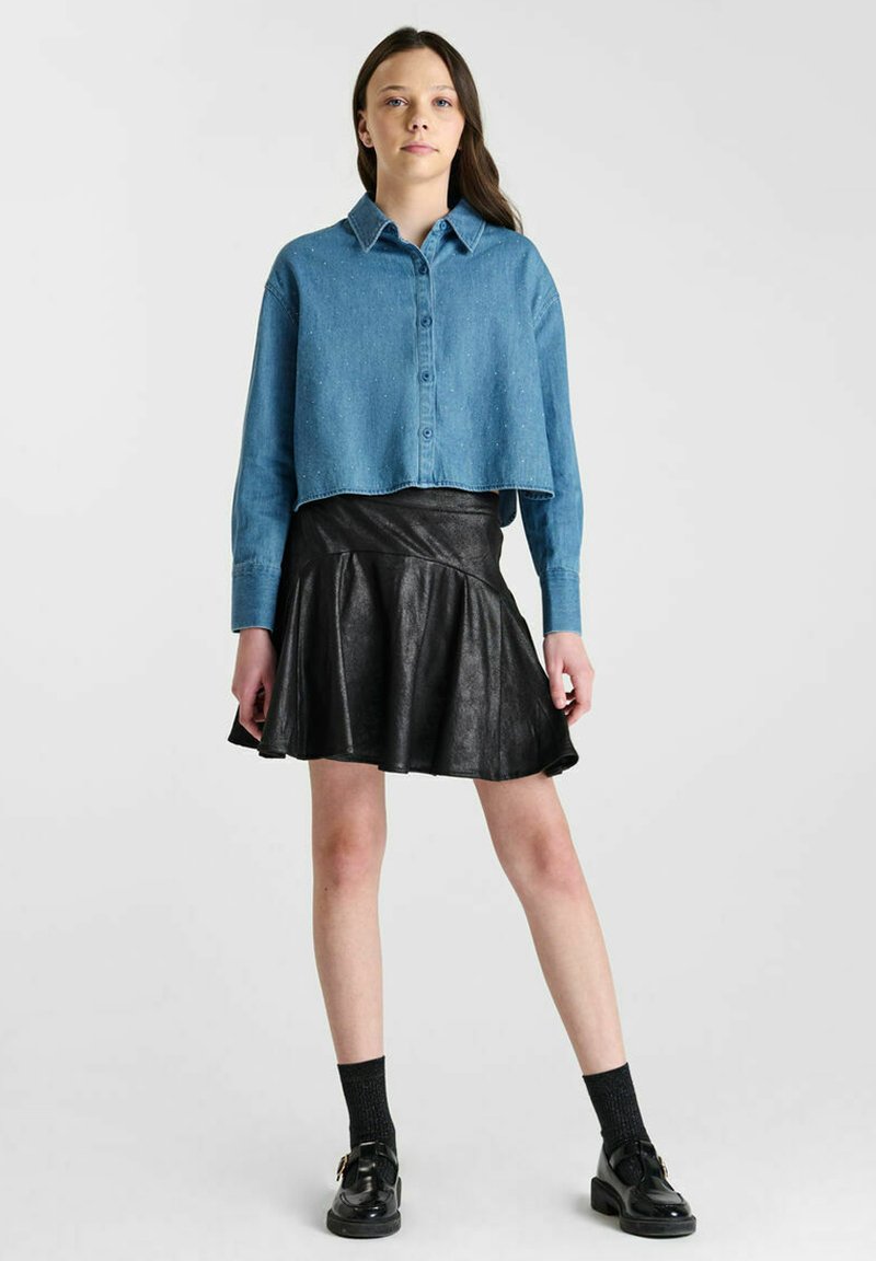 Geknipte blauwe denimshirt met een knopenfront, gecombineerd met een zwarte faux leren flared rok. Het model draagt zwarte sokken en schoenen.