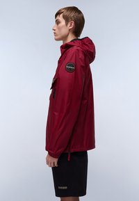 Jong persoon staat zijwaarts en draagt een rode hoodie met logo-opdruk op de mouw en zwarte shorts met logo-opdruk op de zoom.