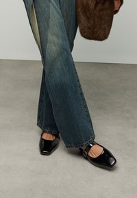 Schwarze Mary-Jane-Schuhe aus Lackleder mit eckiger Zehenpartie, seitlichem Riemen und Schnalle, kombiniert mit weit geschnittenen blauen Jeans.