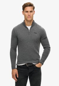 Maglione grigio con zip, maniche lunghe, texture lavorata a maglia e logo ricamato sul lato sinistro del petto. Abbinato a jeans neri e un orologio.