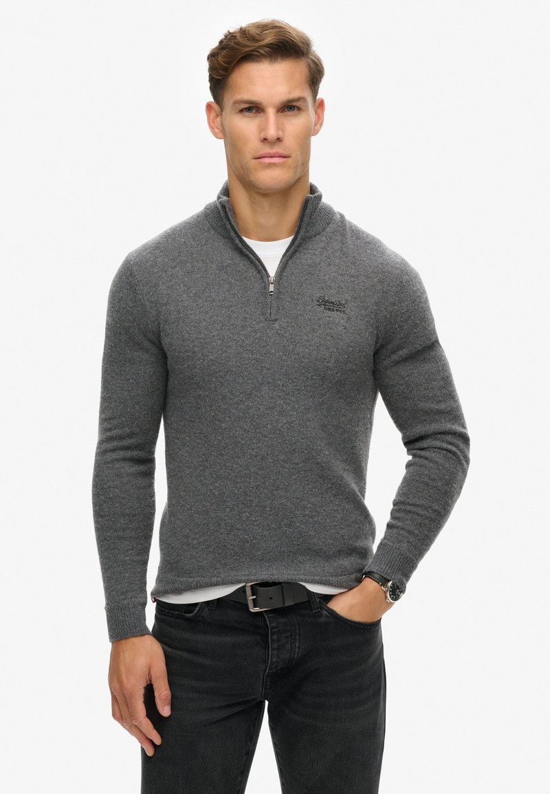 Maglione grigio con zip, maniche lunghe, texture lavorata a maglia e logo ricamato sul lato sinistro del petto. Abbinato a jeans neri e un orologio.