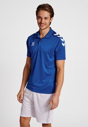 Blå kortærmet polo-shirt i glat stof med krave og hvide chevron-detaljer på skuldrene. Bæres med hvide shorts.