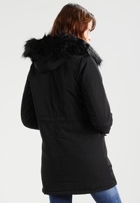 Černá parka s kapucí lemovanou kožešinou, s užším pasem, dlouhými rukávy a hladkým, polstrovaným materiálem.