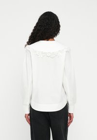 Persona con lunghi capelli ricci, che indossa una blusa bianca con un ampio colletto in pizzo e pantaloni neri, vista di spalle su uno sfondo semplice.