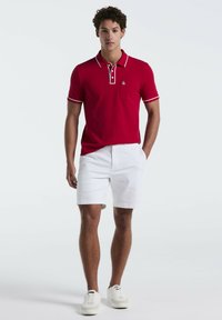 T-shirt polo rouge avec des accents blancs, à manches courtes et une poche poitrine, associé à un short blanc et des chaussures blanches. Coupe simple et sportive.