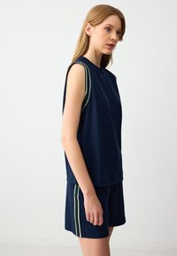 Marineblauw mouwloos topje met uitsnijdingen op de schouders, voorzien van groene en witte gestreepte accenten, gecombineerd met marineblauwe shorts met bijpassende strepen.