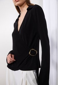 Black long-sleeve wrap top in soft fabric, featuring a deep V-neck and a gold-tone buckle accenting the side. Paired with white pleated pants.