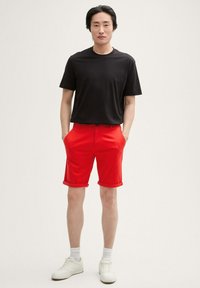 Camiseta negra de manga corta, pantalones cortos rojos arremangados, sin patrones visibles. Zapatillas y calcetines blancos completan el atuendo. Diseño sencillo y colores vibrantes.