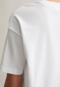 T-shirt blanc à manches courtes avec une texture côtelée et un col ras du cou. Détails de couture visibles, mettant en valeur une coupe décontractée.