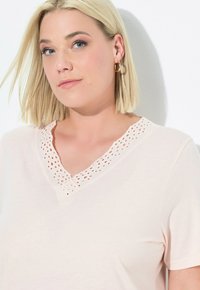 Maglietta rosa chiaro a maniche corte con scollo a V, caratterizzata da dettagli in pizzo occhiellato lungo il colletto e da una texture morbida e liscia.