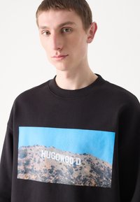 Černá mikina s velkým grafickým zobrazením horské krajiny, na které je bíle napsáno "HUGOWOOD", se pozadím modré oblohy.