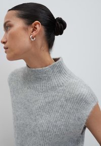Frau mit dunklem Haar im Dutt, die silberne Creolenohrringe und einen ärmellosen hellgrauen gestrickten Rollkragenpullover trägt und nach links schaut.